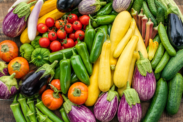 Overhead photo of an abundant fresh vegetable harvest — purple and white eggplants, cherry and heirloom tomatoes, green chile peppers, yellow squash, okra, and cucumbers — arranged on a rustic wood surface.